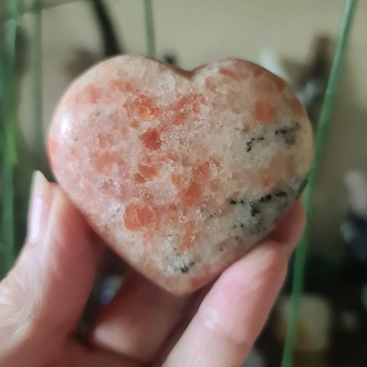A red polished heart-shaped sunstone crystal being held in a person's hand.