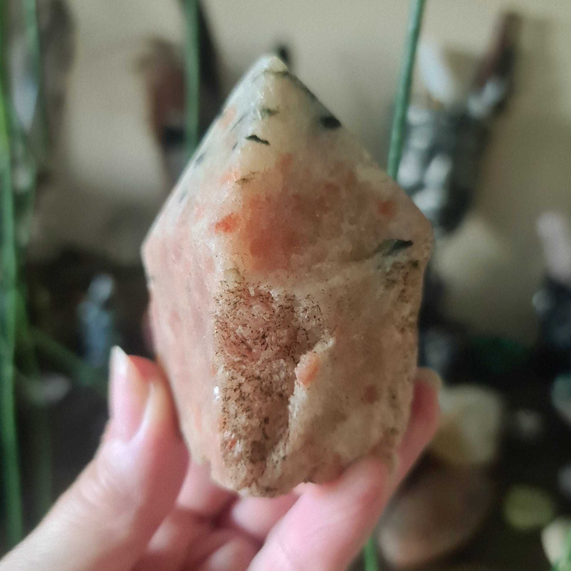 A polished red sunstone point held in a person's hand, with a blurred background of various crystal figures and objects.