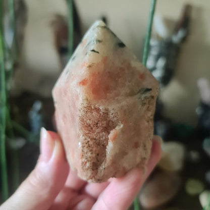 A polished red sunstone point held in a person's hand, with a blurred background of various crystal figures and objects.