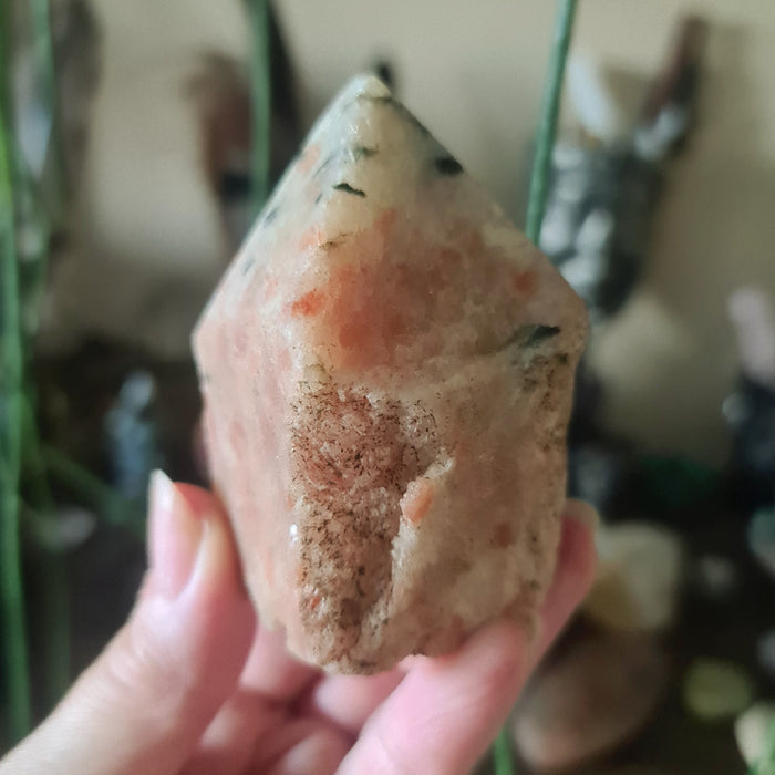 A polished red sunstone point held in a person's hand, with a blurred background of various crystal figures and objects.