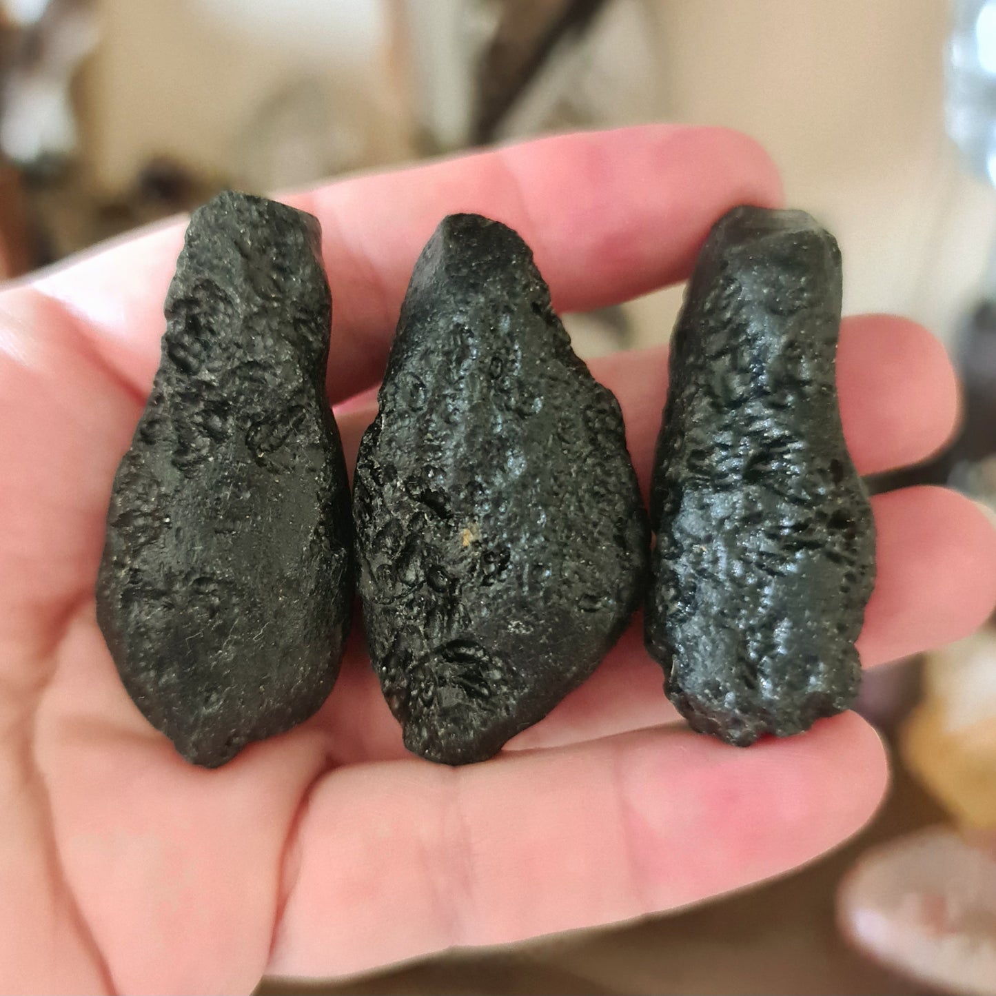 Three black tektite teardrop-shaped stones held in a person's hand, with a blurred background.