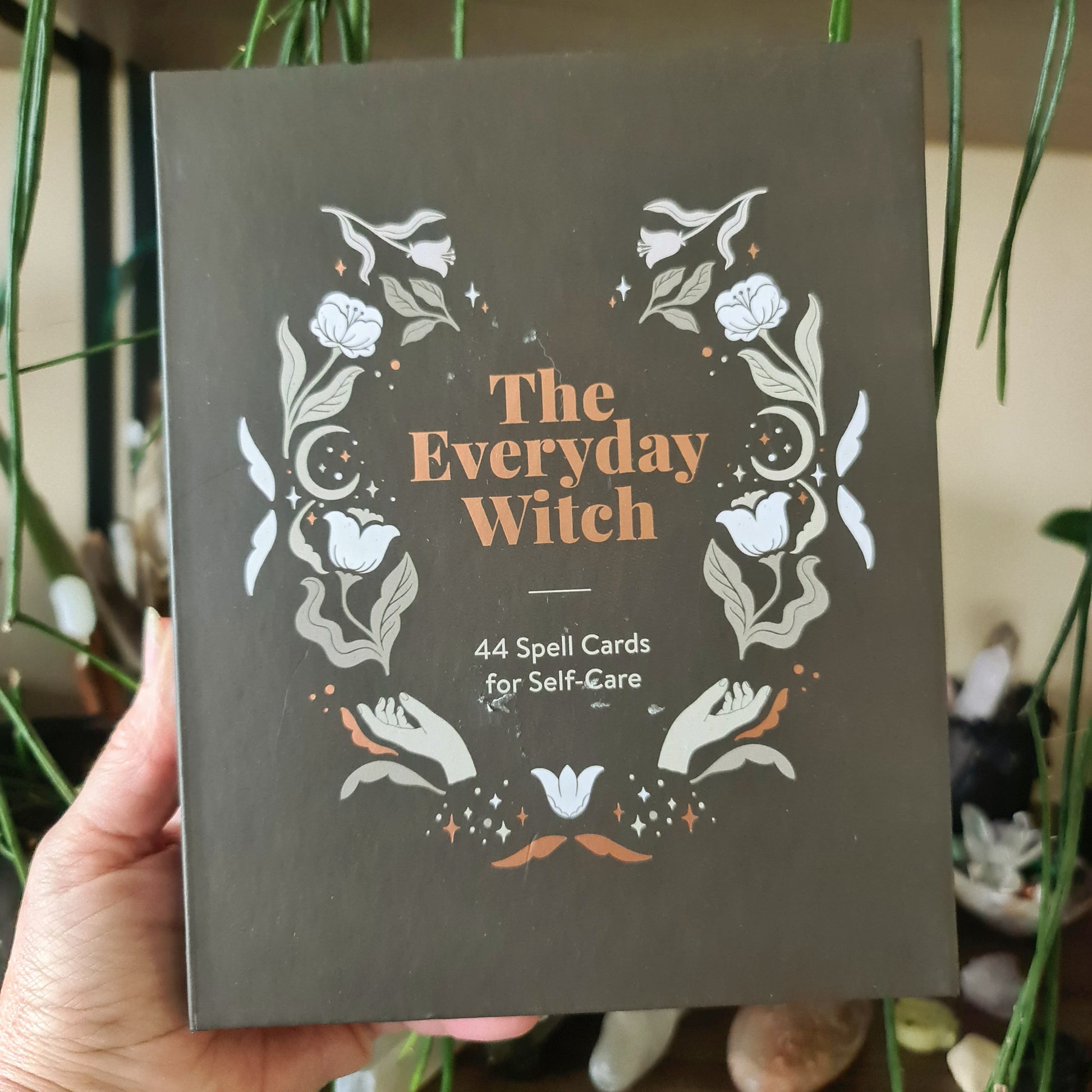 A boxed set titled 'The Everyday Witch' containing 44 spell cards for self-care, held in a person's hand, with a design of herbs and flowers on the cover.