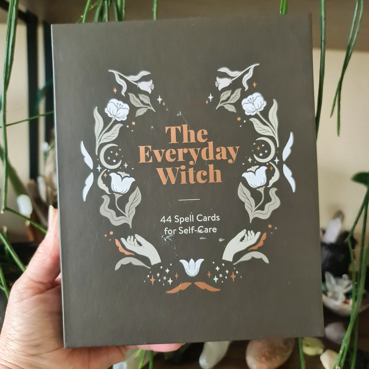A boxed set titled 'The Everyday Witch' containing 44 spell cards for self-care, held in a person's hand, with a design of herbs and flowers on the cover.