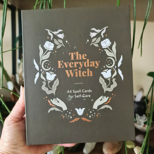 A boxed set titled 'The Everyday Witch' containing 44 spell cards for self-care, held in a person's hand, with a design of herbs and flowers on the cover.