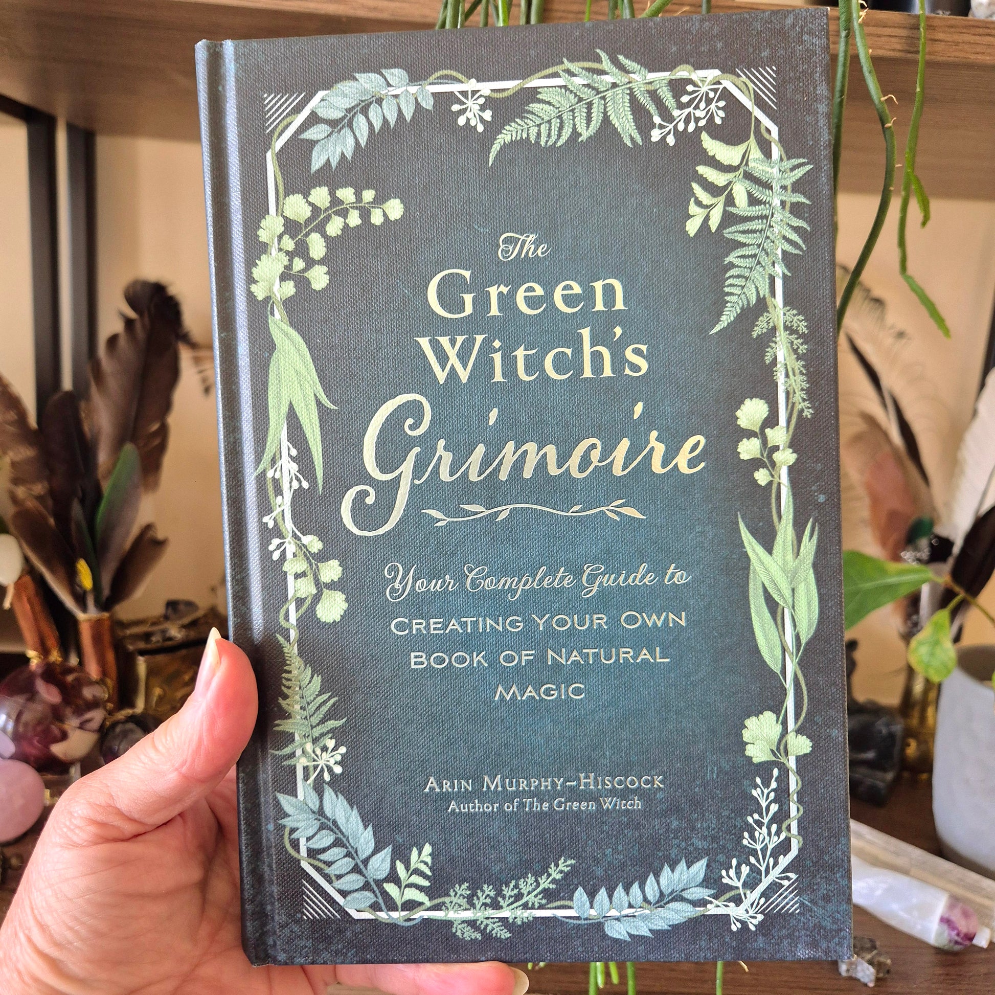 A hand holding a book titled 'The Green Witch's Grimoire' by A.M.