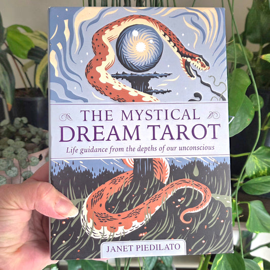 A hand holds a boxed tarot card deck titled "The Mystical Dream Tarot" by Janet Piedilato. The box art shows a stylized snake wrapping around a glowing orb against a blue sky and dark forest background.
