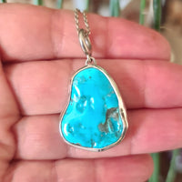 A sterling silver pendant with a turquoise stone held in a hand, with a plant in the background.