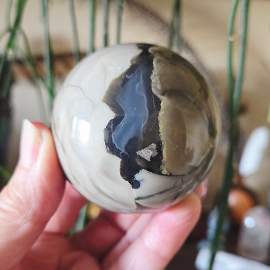 A hand holding a Volcano Agate Sphere with a pattern of predominantly gray and black colors.