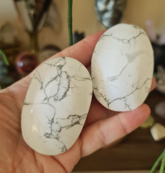 Two polished White Howlite Palm Stones held in a person's hand, displaying a marbled white and gray pattern.