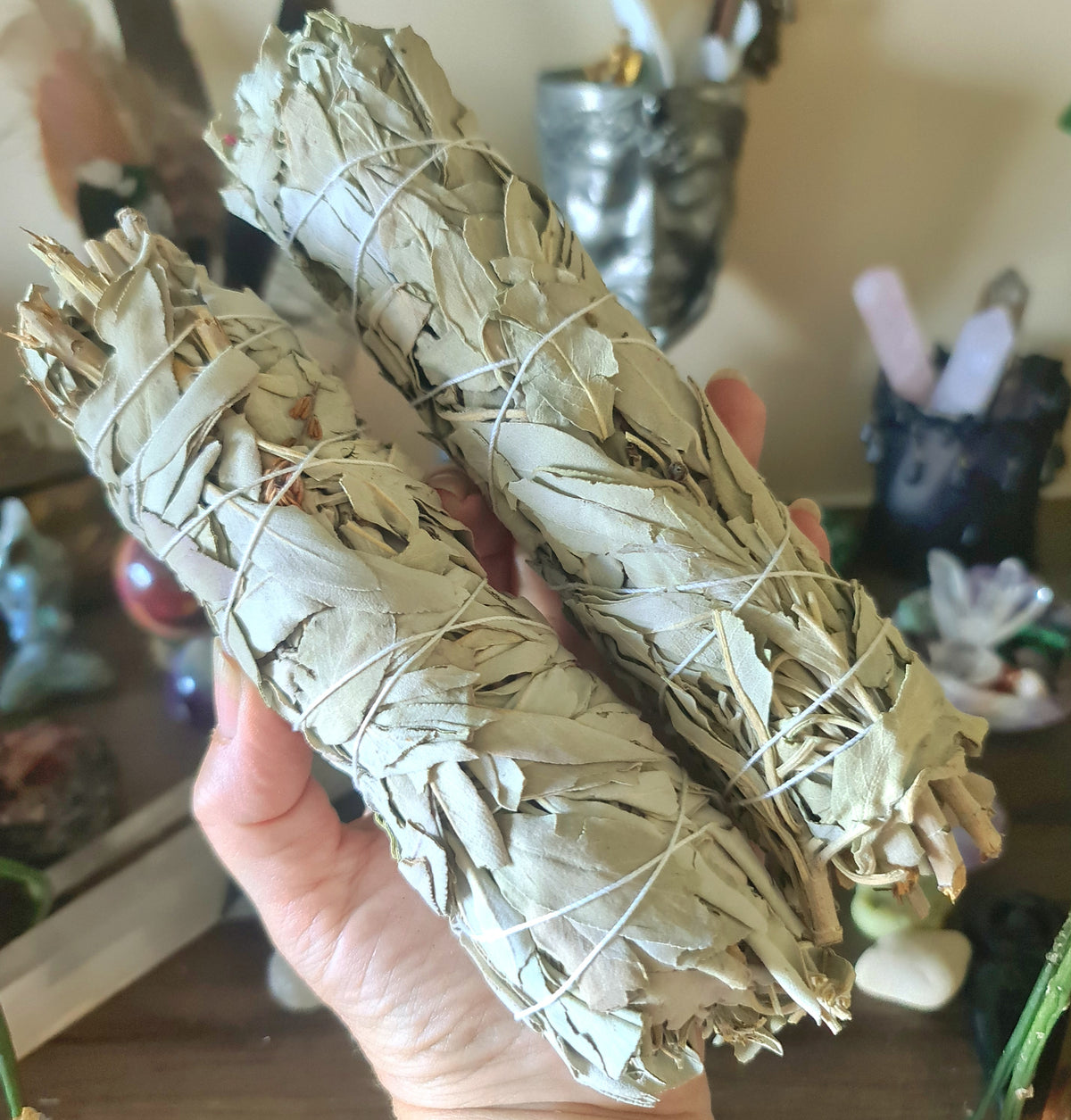 A hand holding a large sage smudge stick with string, in a ceremonial bundle format, with various crystal figures and items in the background.
