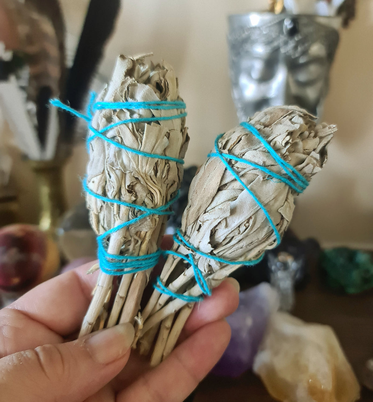 Two white sage smudge sticks tied with blue ribbons and a silver knife, held in hands, with various spiritual items in the background.