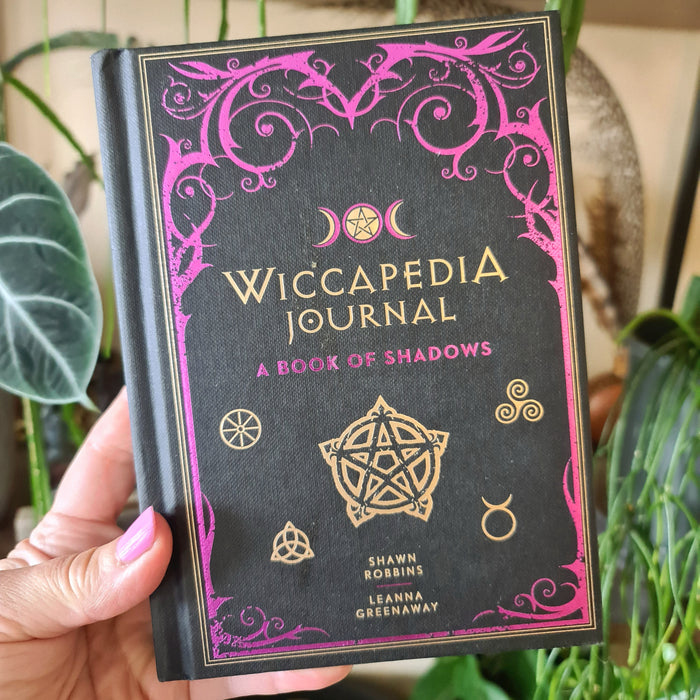 A hand holding a book titled 'Wiccapedia Journal - A Book of Shadows' with a pink and black cover featuring a pentagram and other symbols.