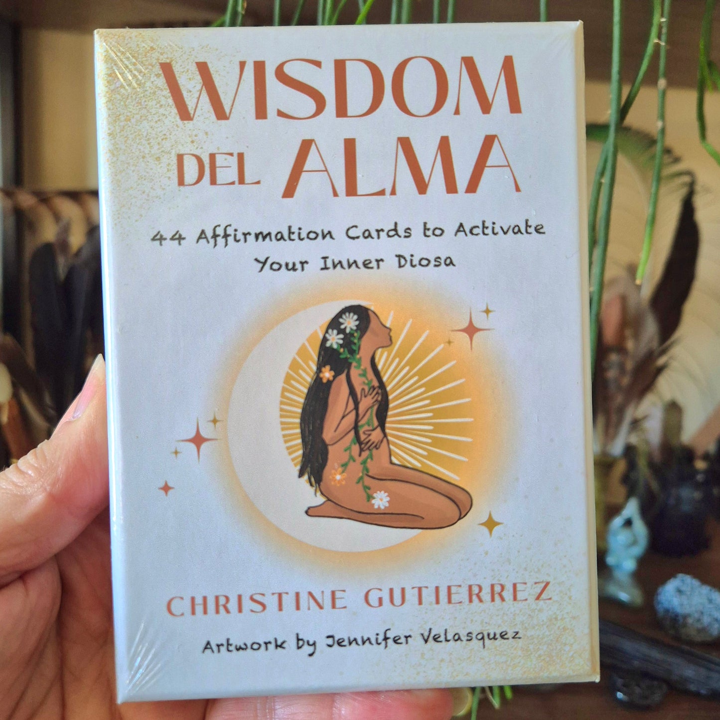 A book titled 'Wisdom Del Alma' with a subtitle '4 Affirmation Cards to Activate Your Inner Diva' held in a hand. The cover features an illustration of a woman sitting under a sun with flowers in her hair, surrounded by decorative stars and the word 'WISDOM' at the top.