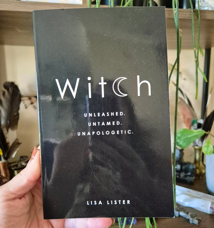 This image features a person holding a book titled "Witch" by Lisa Lister. The book's cover is strikingly simple and elegant, primarily in black with a white band that carries the title in a stylish, scripted font.