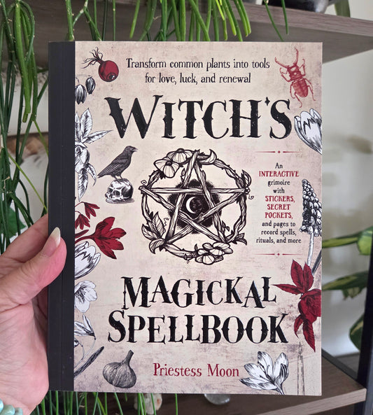 A hand holds a hardcover book titled "Witch's Magickal Spellbook" by Priestess Moon. The cover has a beige parchment-style background with black text, a central pentagram illustration, and red and black botanical and animal drawings.