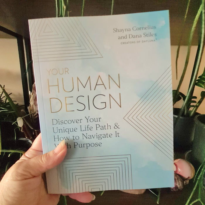 A book titled 'Your Human Design' by Shayna Correla-Stern and Dana Stiles, published by DayLuna, is held in hand, with a blue and white cover design featuring geometric shapes.