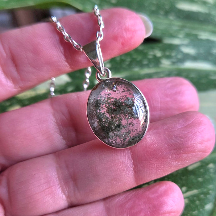 Silver necklace with a round pendant held in a hand against a green leafy background