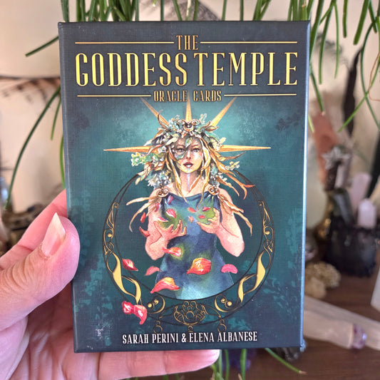 Book titled 'The Goddess Temple Oracle Cards' held by a hand with plants in the background