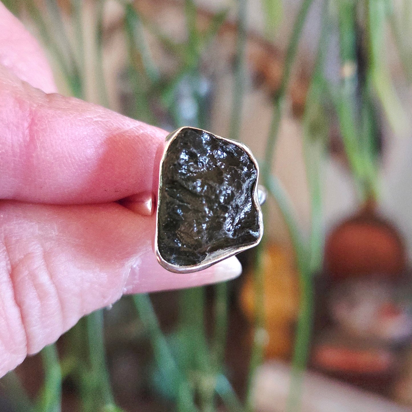 Hand holding a ring with a large, rough-cut gemstone against a blurred natural background