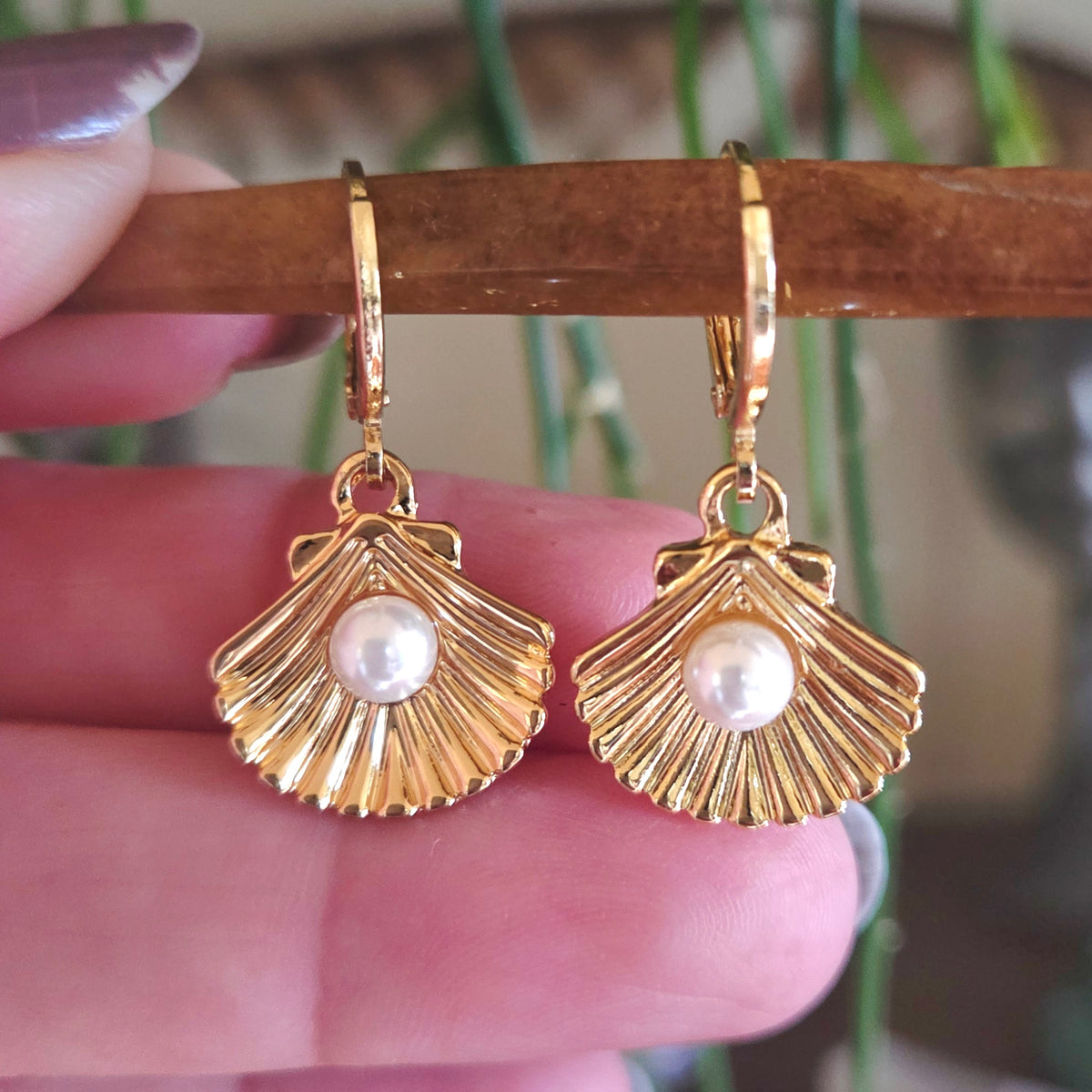 Gold shell-shaped earrings with pearl centers held by a hand against a blurred background.
