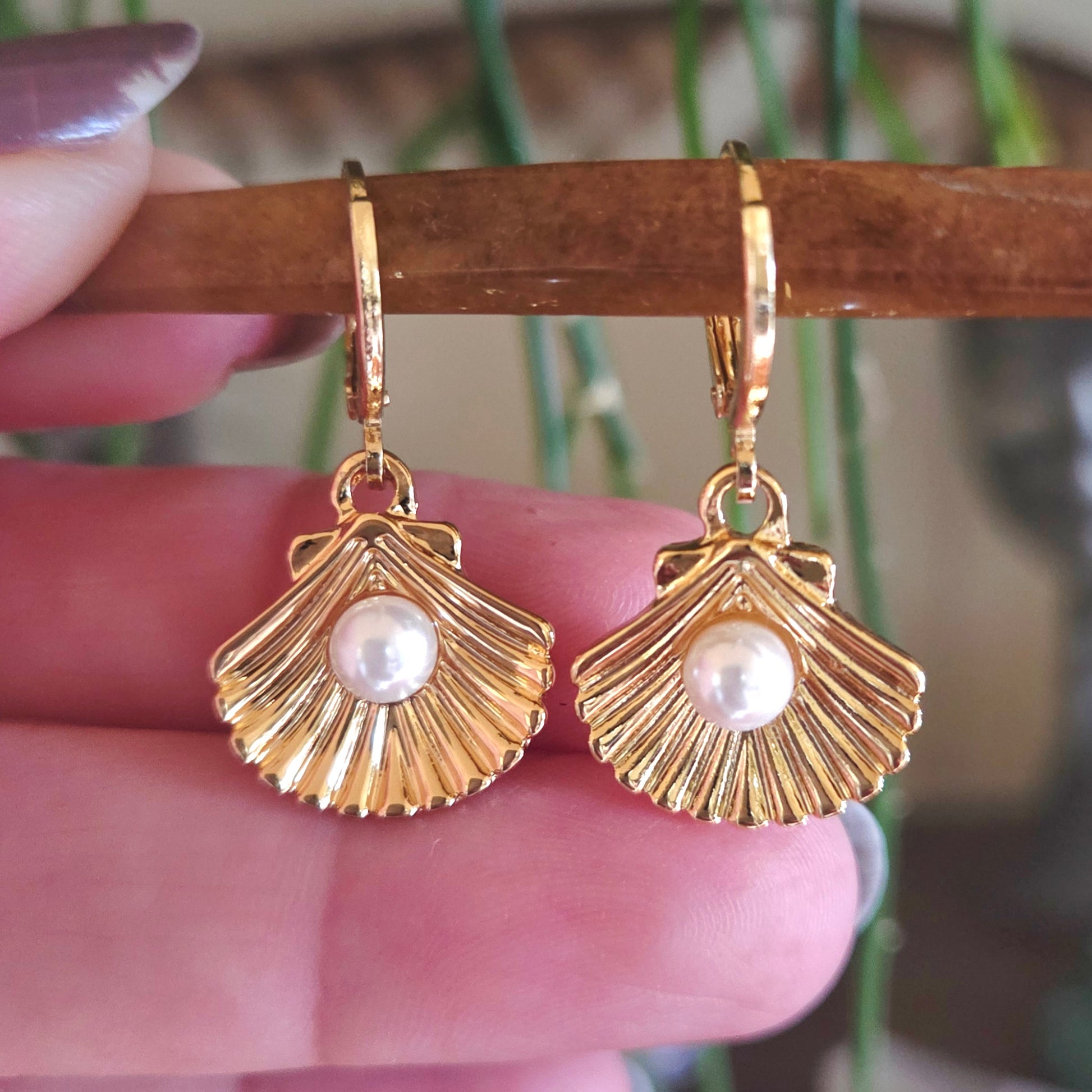 Gold shell-shaped earrings with pearl centers held by a hand against a blurred background.