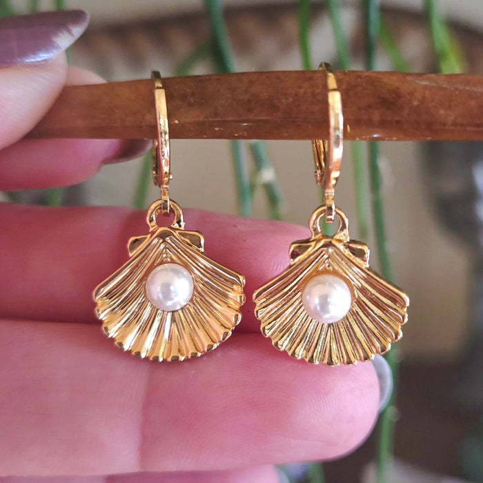 Gold shell-shaped earrings with pearl centers held by a hand against a blurred background.