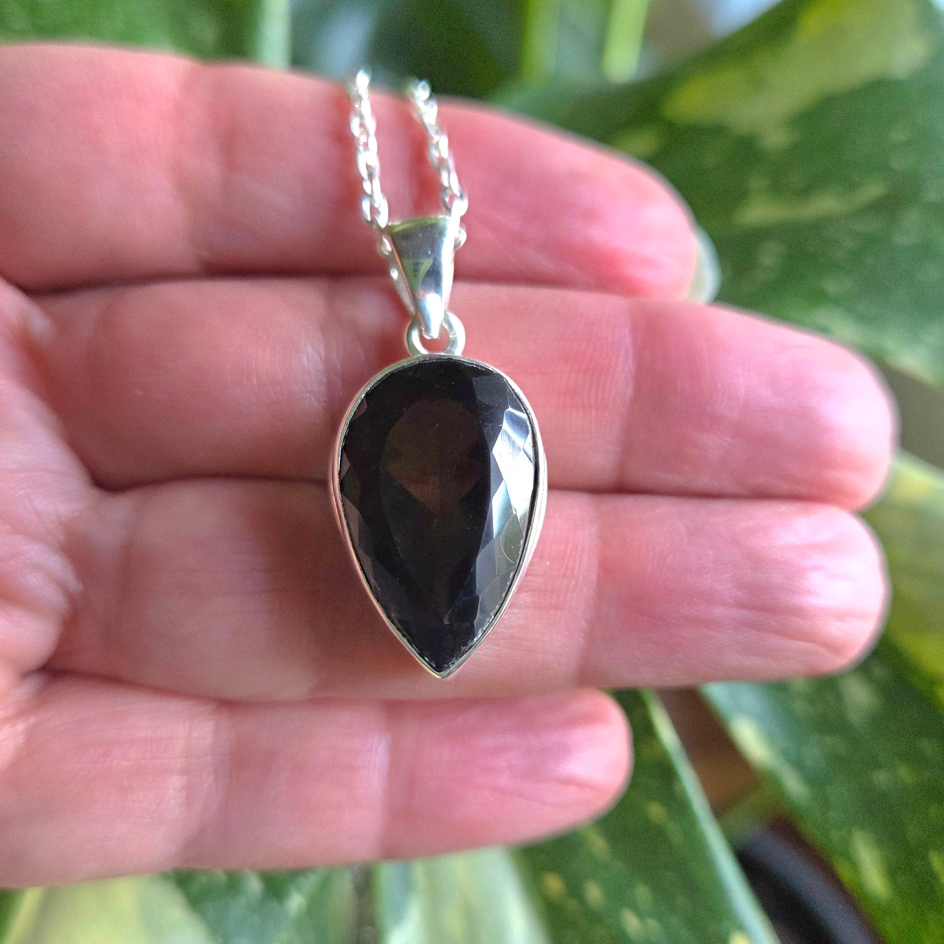 Teardrop-shaped transparent stone pendant on a silver chain held in a hand with a green leafy background.