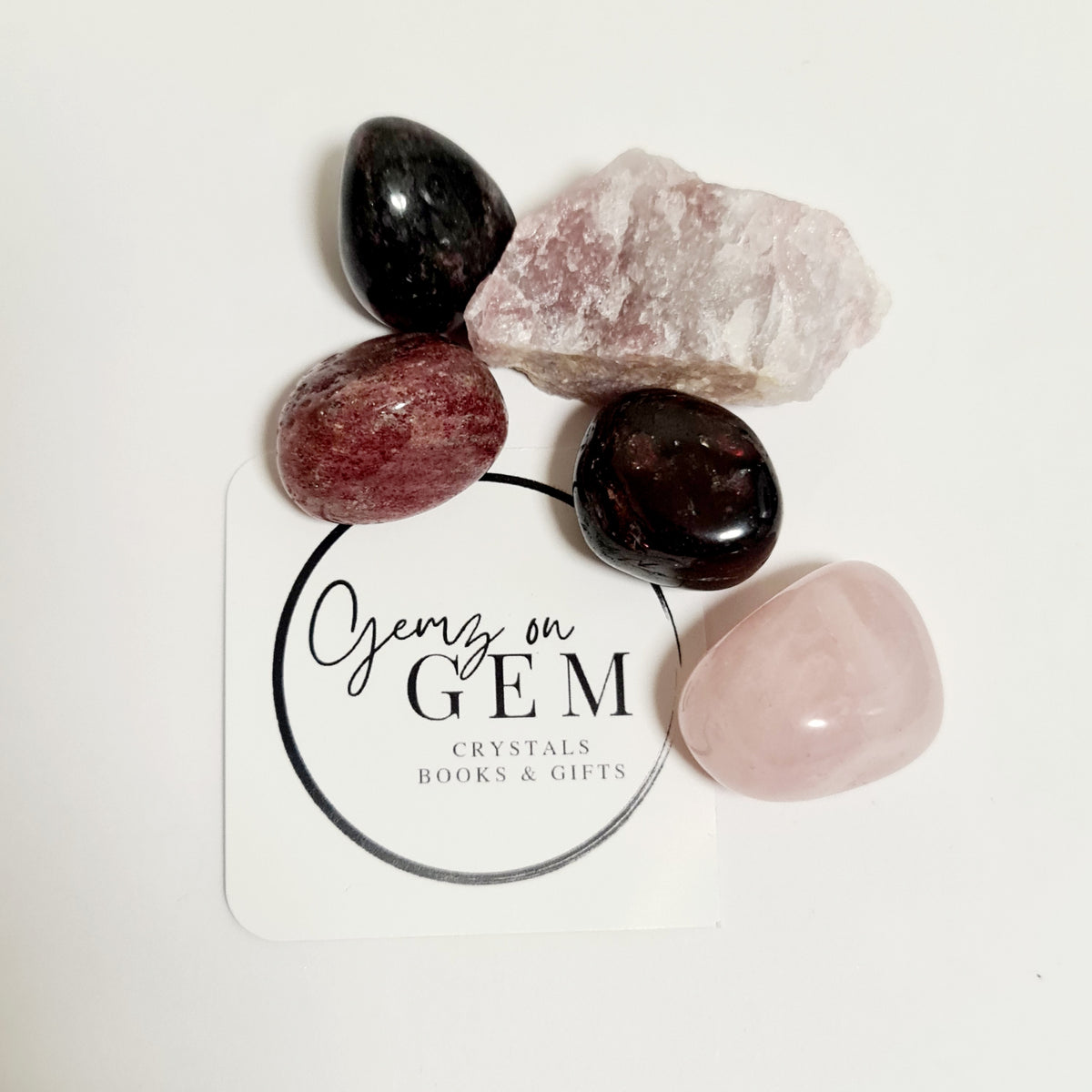 A collection of polished gemstones including a pink pearl, red agate, black onyx, and pink quartz, arranged on a surface with a card that reads 'Gemz on Gem'.
