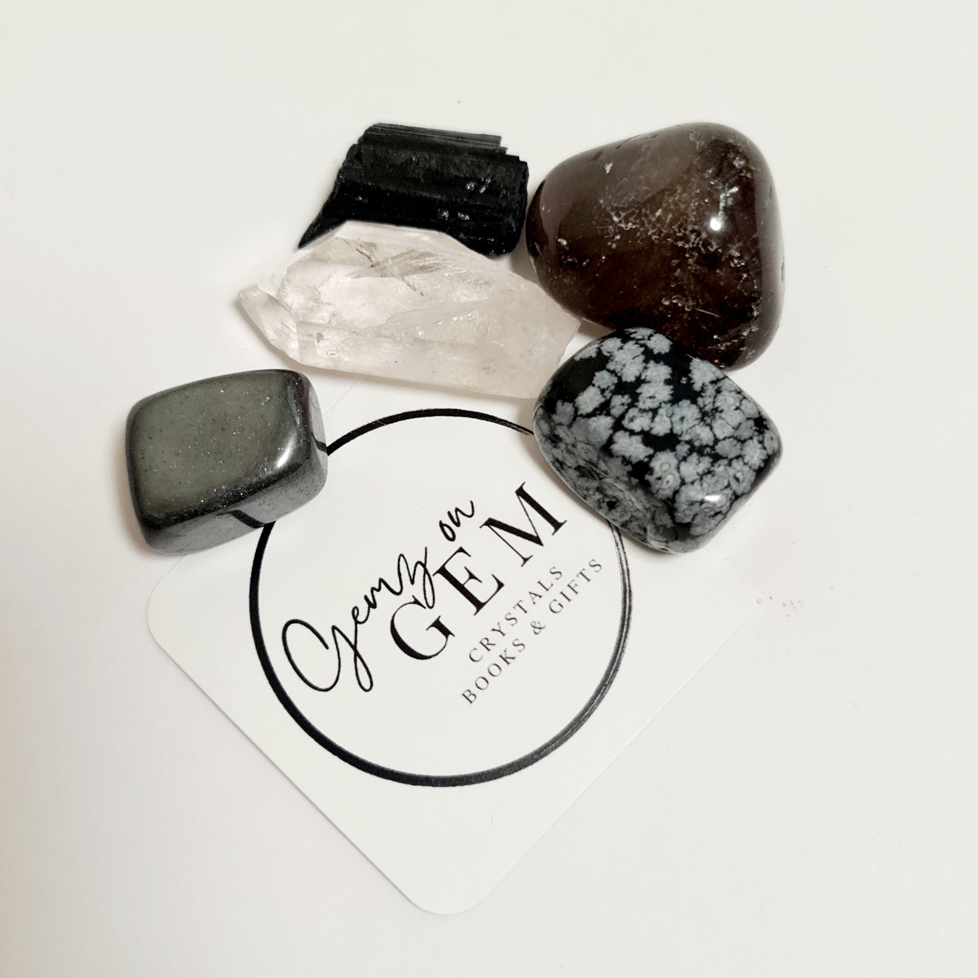 An assortment of polished crystals including black tourmaline, smoky quartz, hematite, and snowflake obsidian, displayed on a branded card with 'Gemz on Gem' printed on it.
