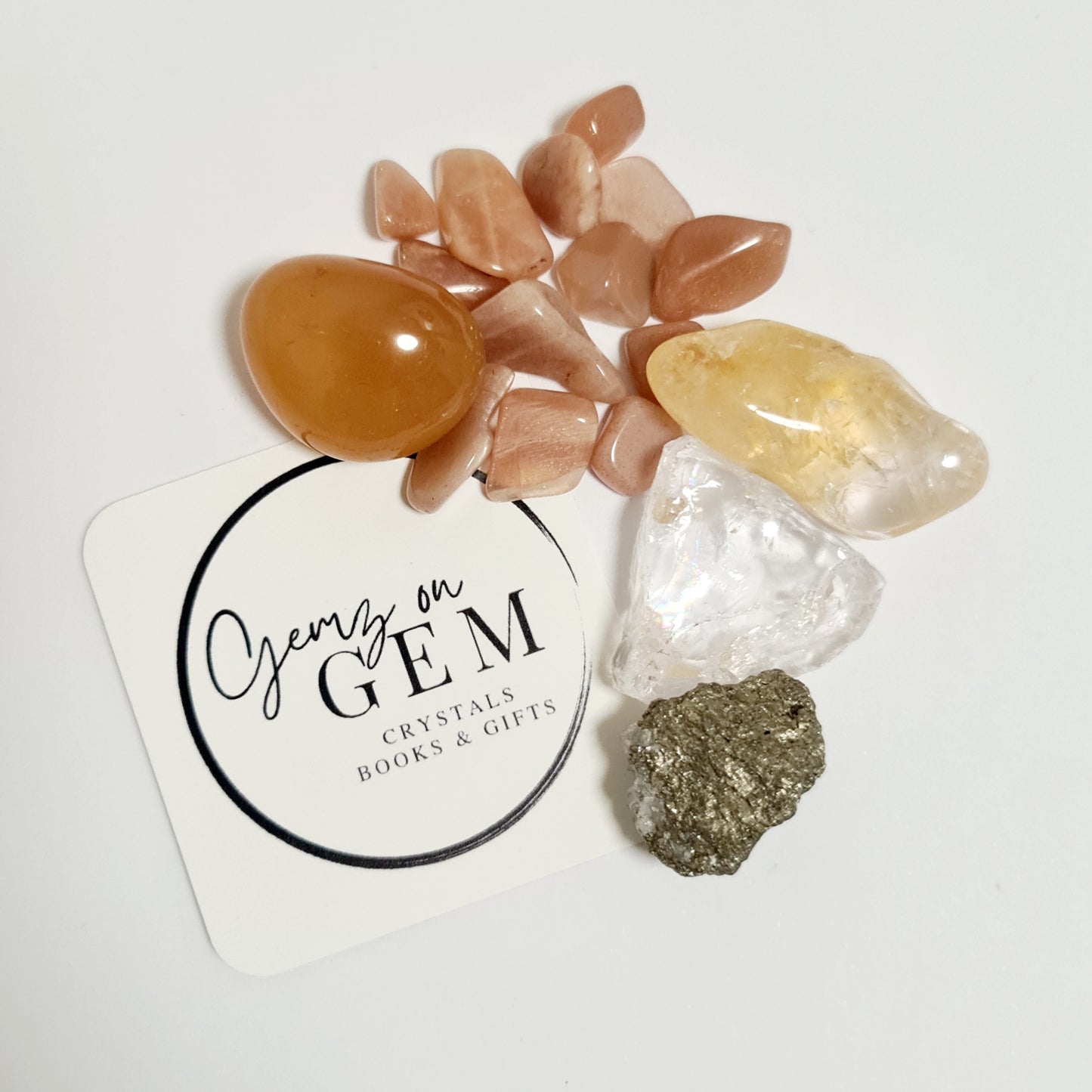 A collection of polished and unpolished crystals including a carnelian, a citrine, a peach moonstone, a pyrite, and a clear quartz, arranged on a white surface with a card that reads 'Crystals on Gem'.