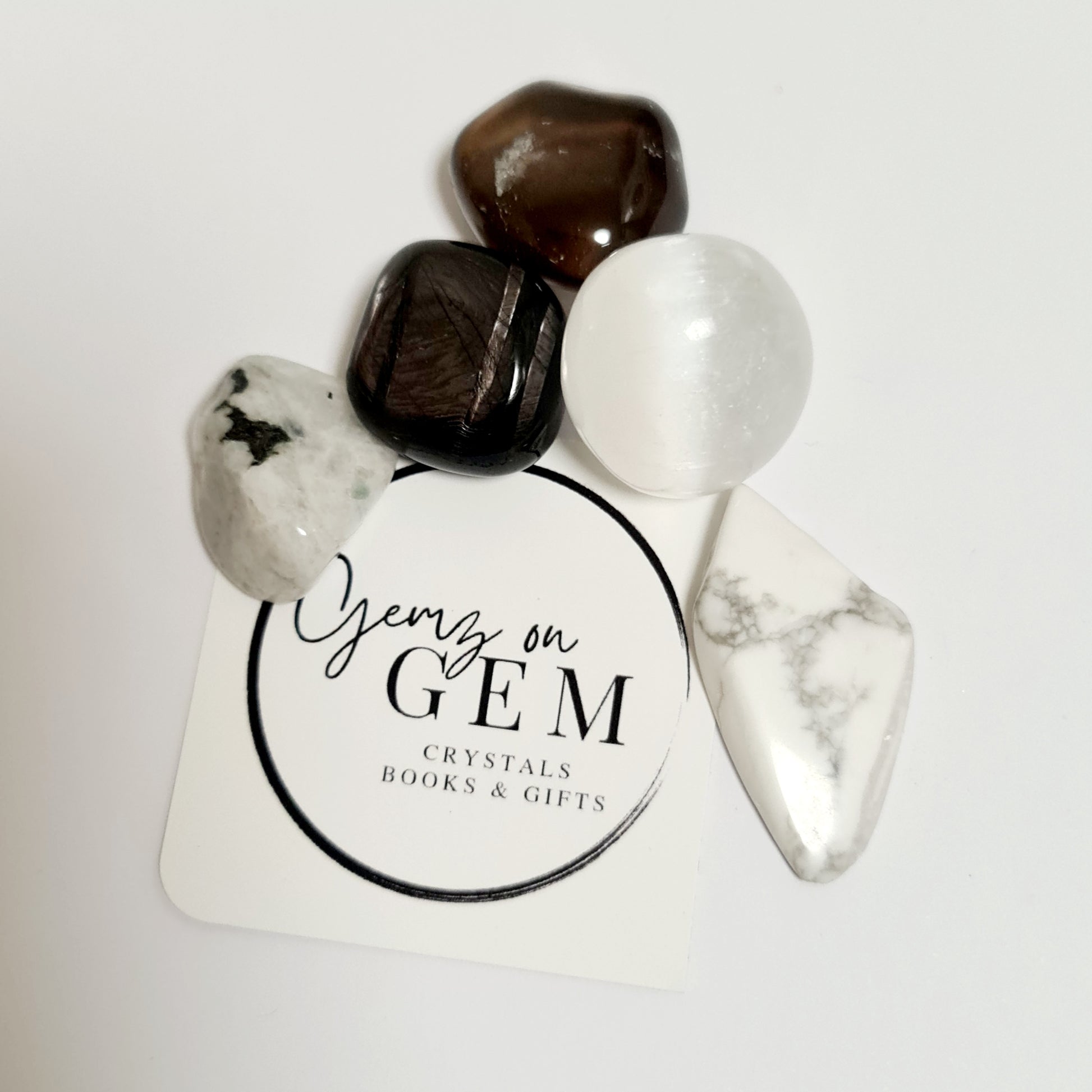An arrangement of various polished crystals including selenite, howlite, hypersthene, rainbow moonstone, and smoky quartz on a branded card with 'Gemz on Gem' printed on it.